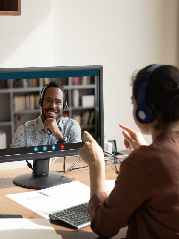 Rear back view Indian woman sit at workplace desk talk to African colleague use pc and video call app. Educational webinar participants, negotiations remotely. Online class teacher and learner concept